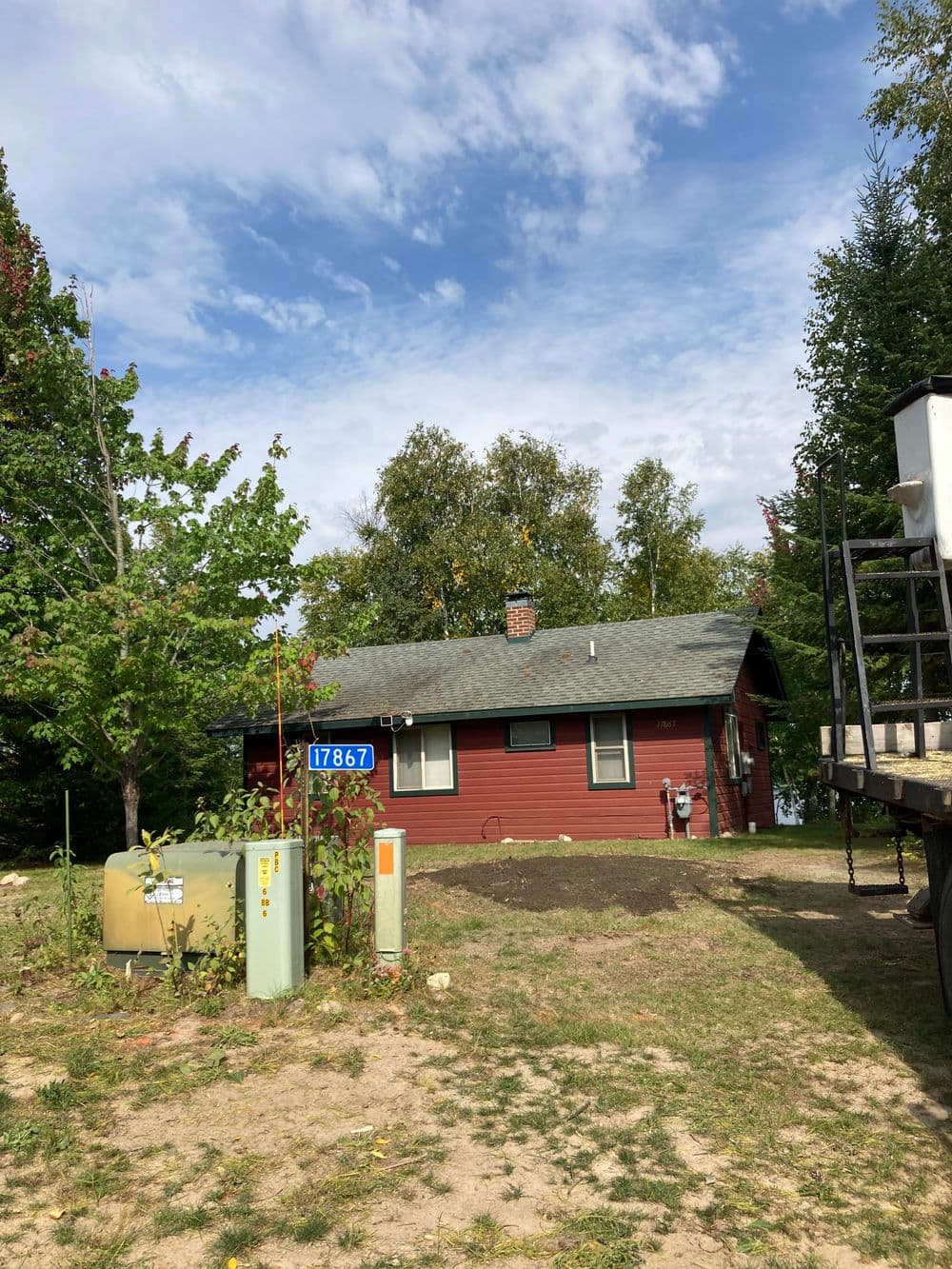 Red house with a gray roof surrounded by trees and utility equipment, under a cloudy sky.