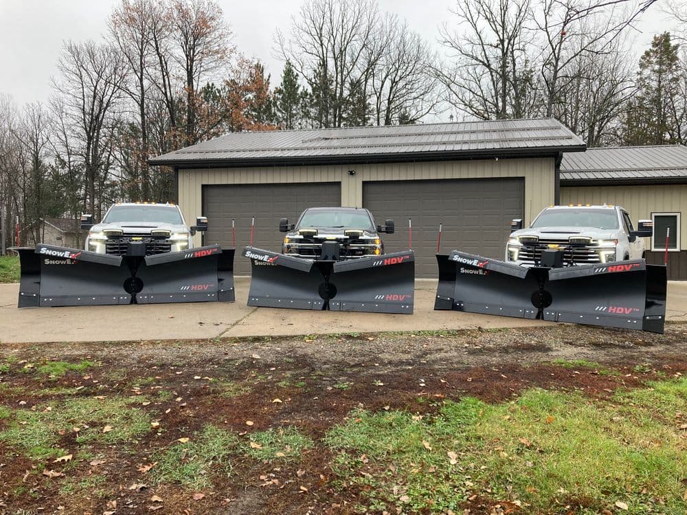 Three snow plows mounted on trucks in front of a garage, surrounded by trees.