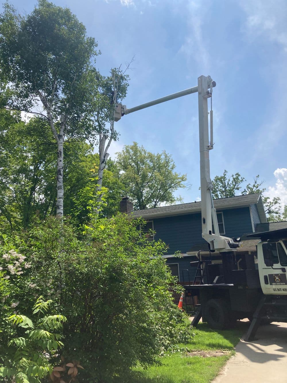 Tree trimming service using a cherry picker near a home with foliage and clear blue sky.
