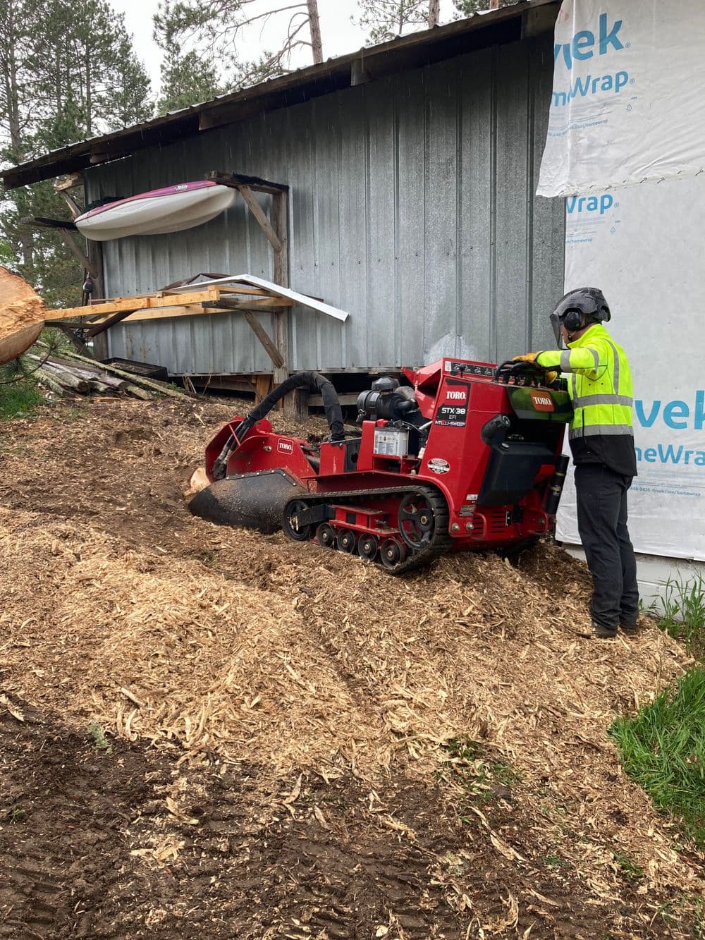 Tree stump grinder in action near a building, with operator wearing safety gear.