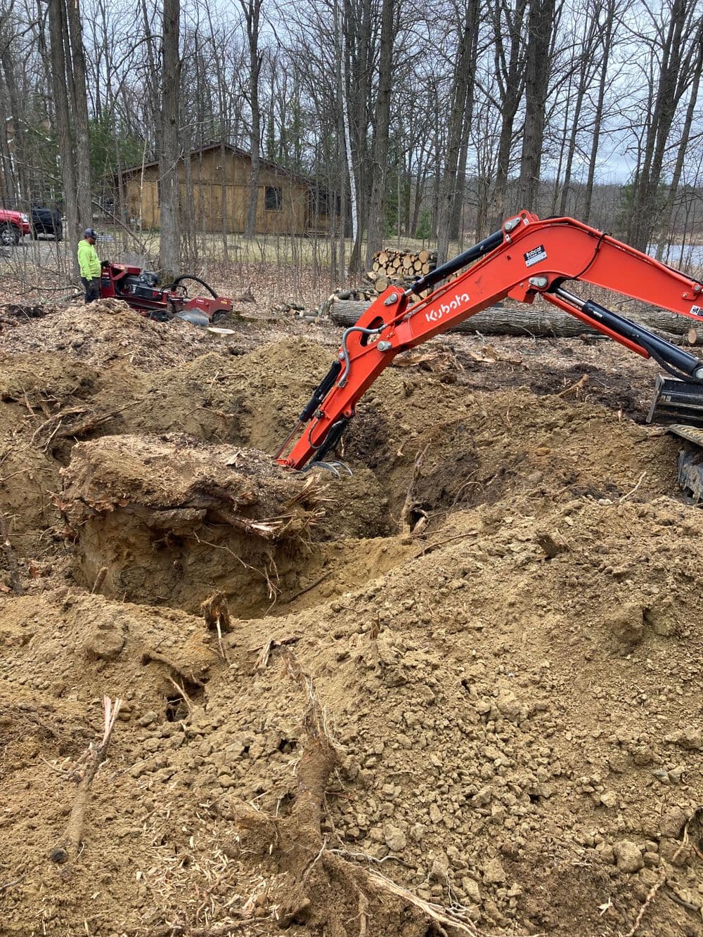 Excavator digging a trench in a wooded area with heavy machinery nearby.
