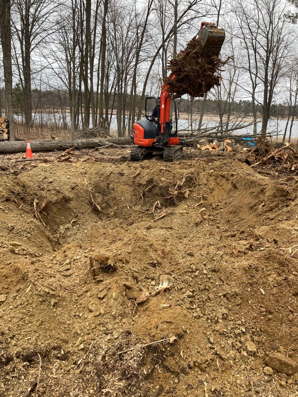 Excavator lifting debris from a large dug hole near a wooded area by a lake.