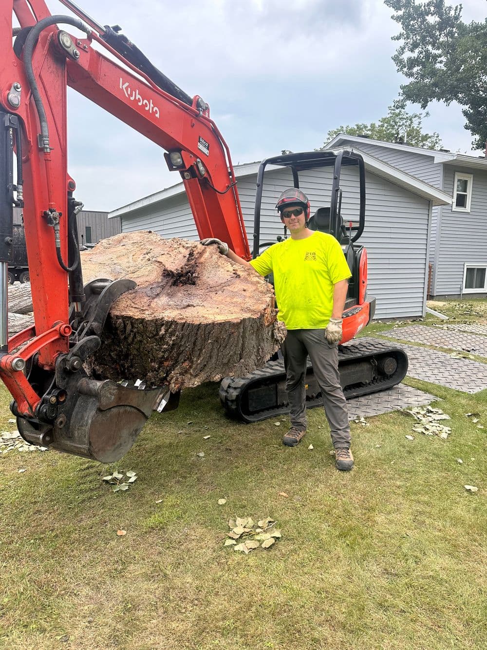 Tree removal expert using an excavator to lift a large log in a residential yard.