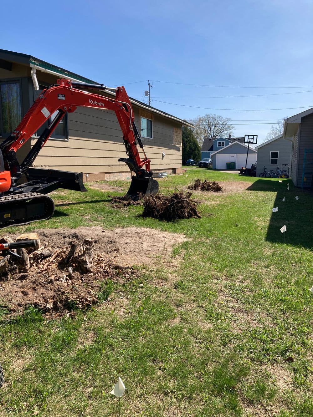 Excavator on residential lawn, preparing ground with tree stump removal and clear blue sky.